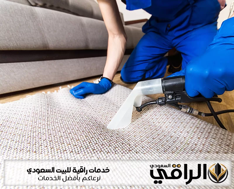 عامل من شركة تنظيف مفروشات بتبوك بزي وقفازات زرقاء ينظف سجادة فاتحة بفوهة شفط أمام كنب رمادي في منزل