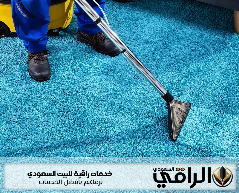 عامل شركة تنظيف مفروشات بتبوك ينظف سجادة زرقاء كثيفة الوبر بفوهة شفط معدنية يظهر شعار الراقي السعودي