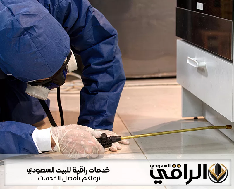 فني شركة مكافحة الحشرات بتيماء يرتدي ملابس واقية ويرش المبيد أسفل خزائن المطبخ لمعالجة أماكن الاختباء